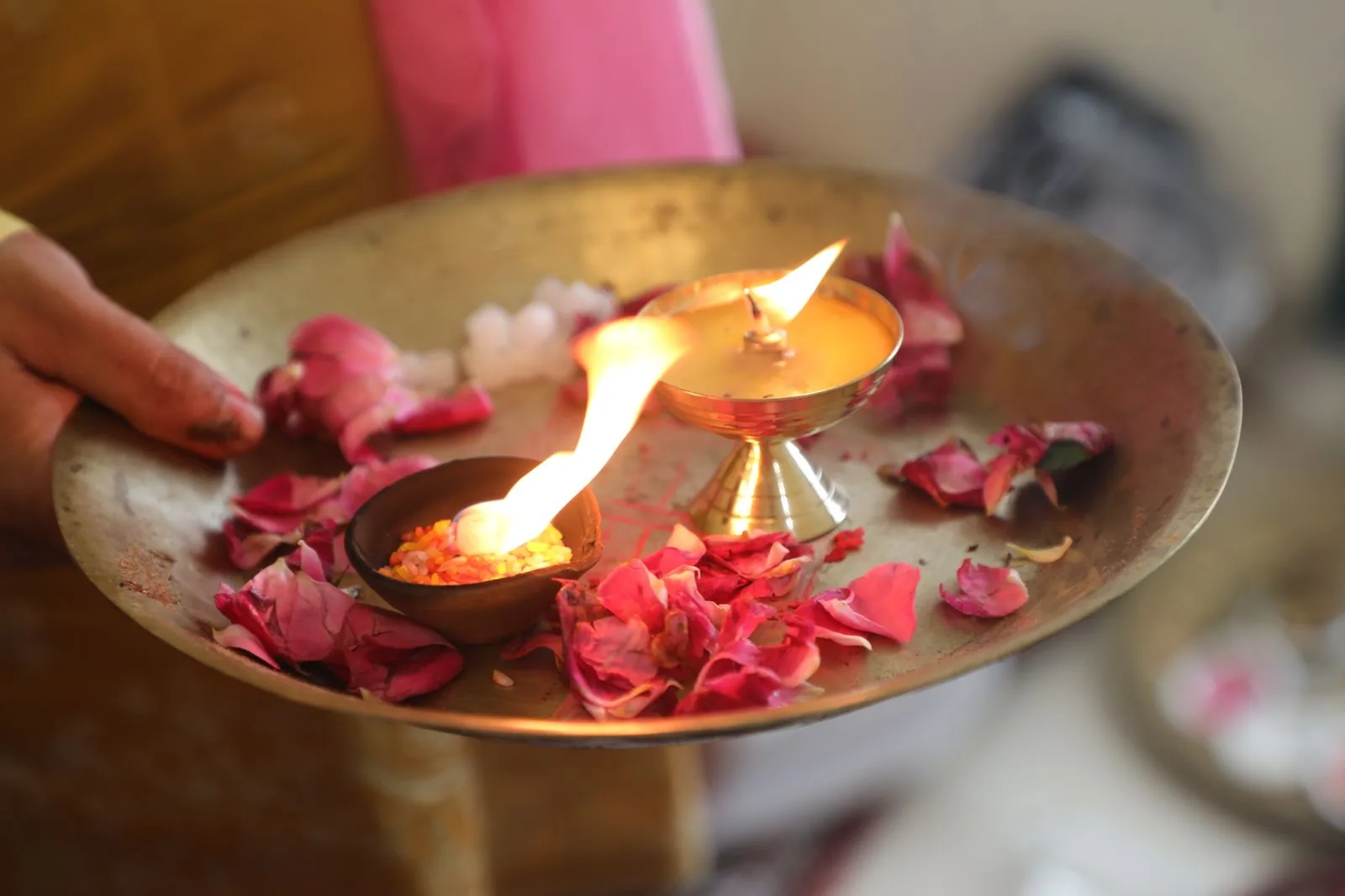 Brass aarti plate with a lit diya and rose petals held in a pilgrim's hand