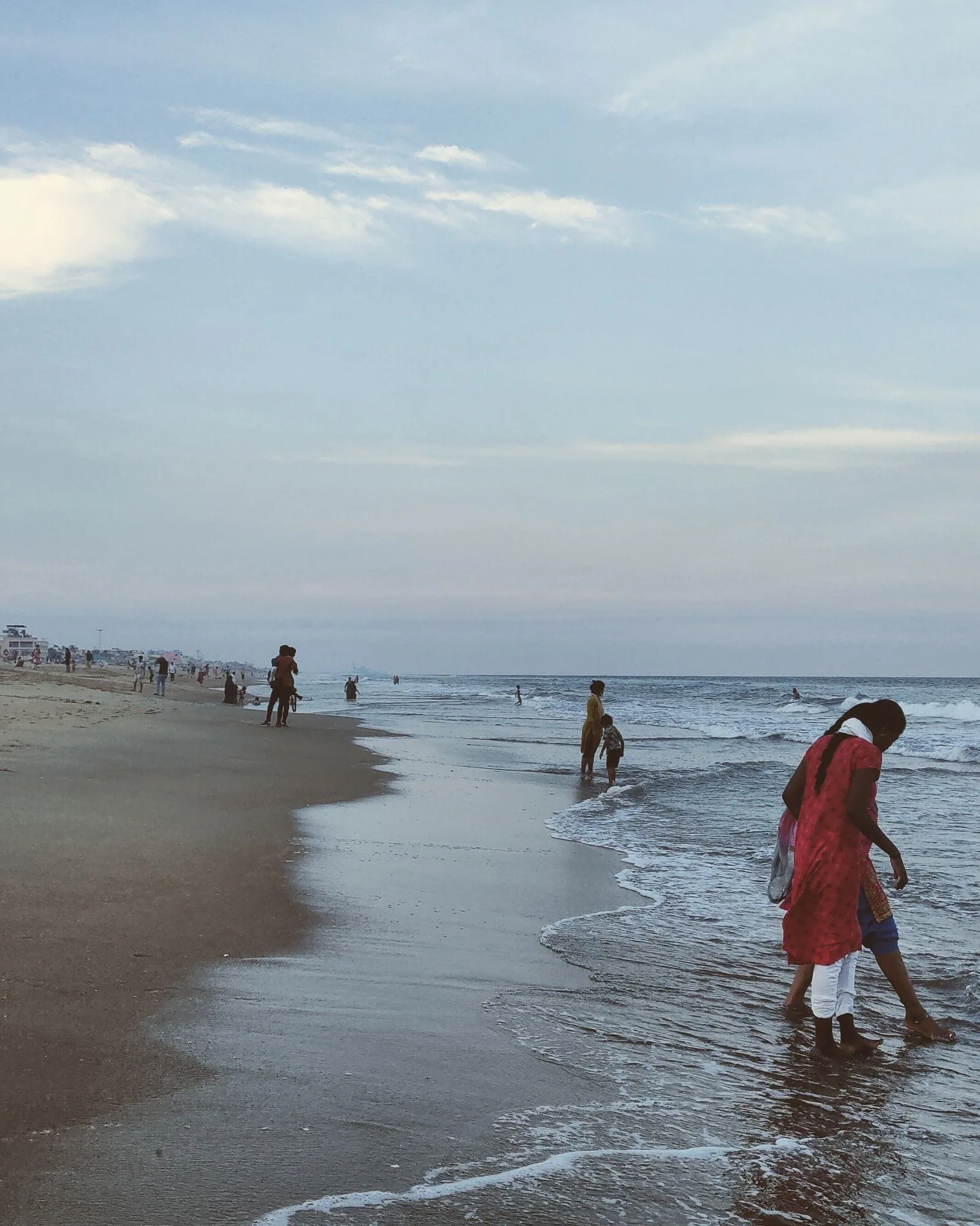 A woman in a red sari wading at the water's edge on a wide sandy beach, others walking along the shore