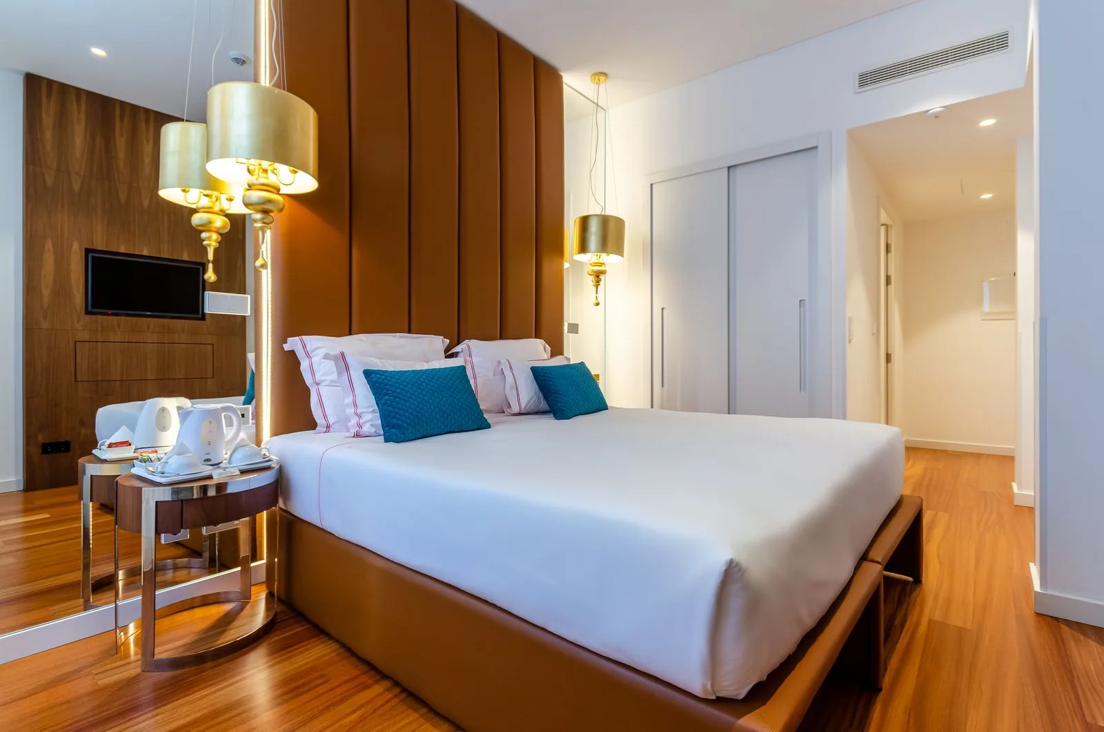 Bright guest room with a made double bed, white linen, teal cushions and a warm wooden floor